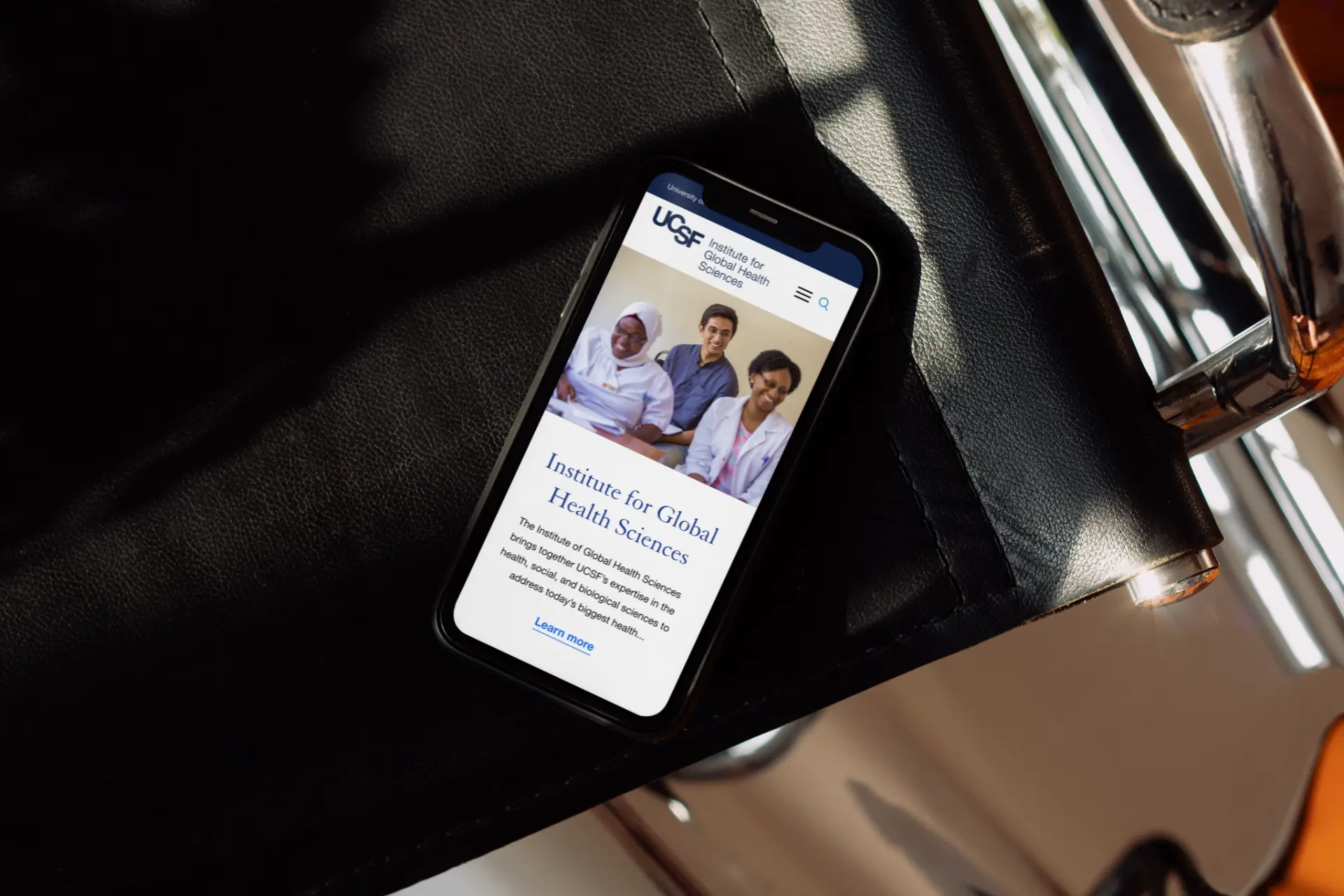 UCSF IGHS mobile homepage shown on a phone resting on a leather seat.