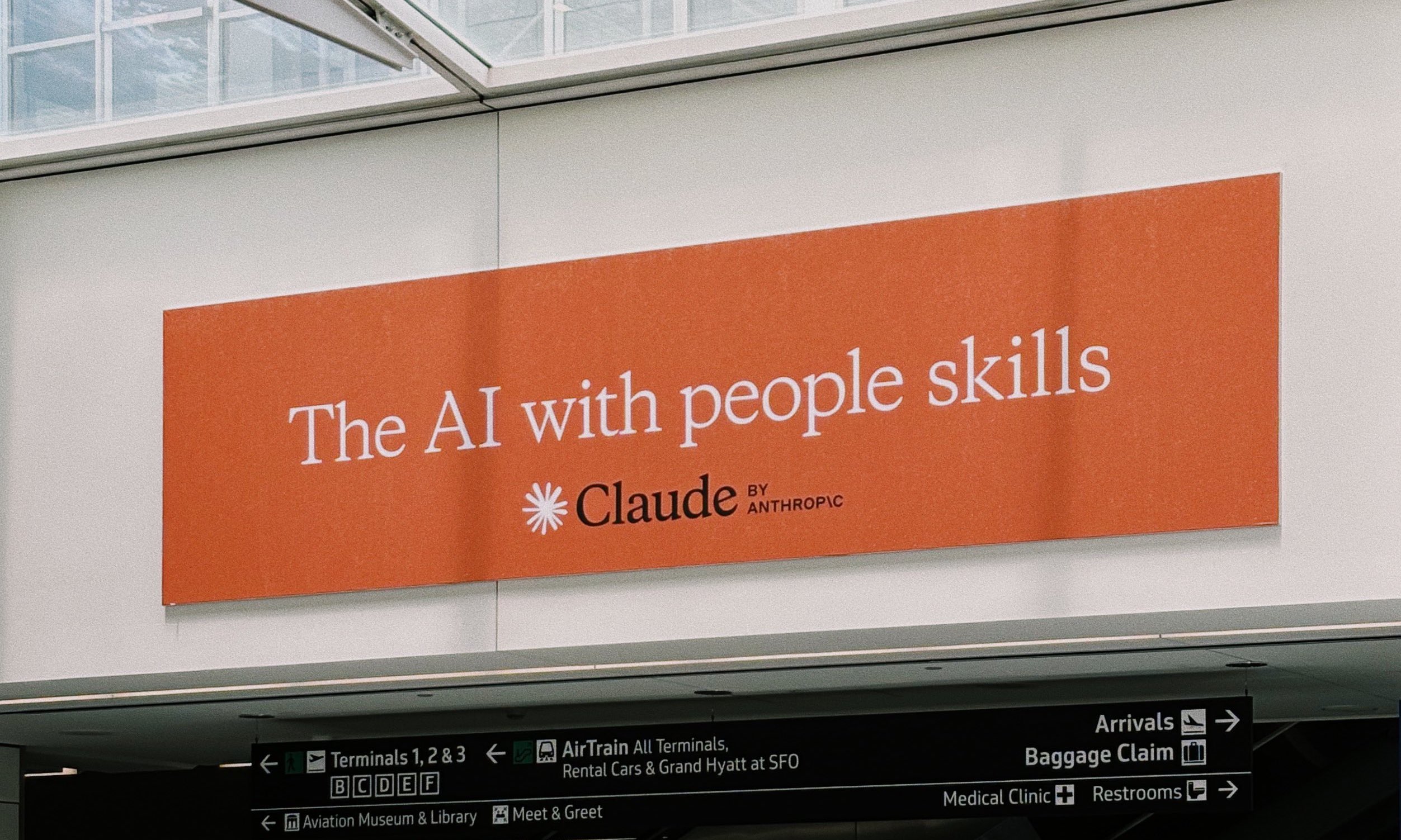 AI design advertisement at SFO airport featuring an Anthropic Claude billboard in coral orange with white text reading 'The AI with people skills', displayed above terminal wayfinding signage